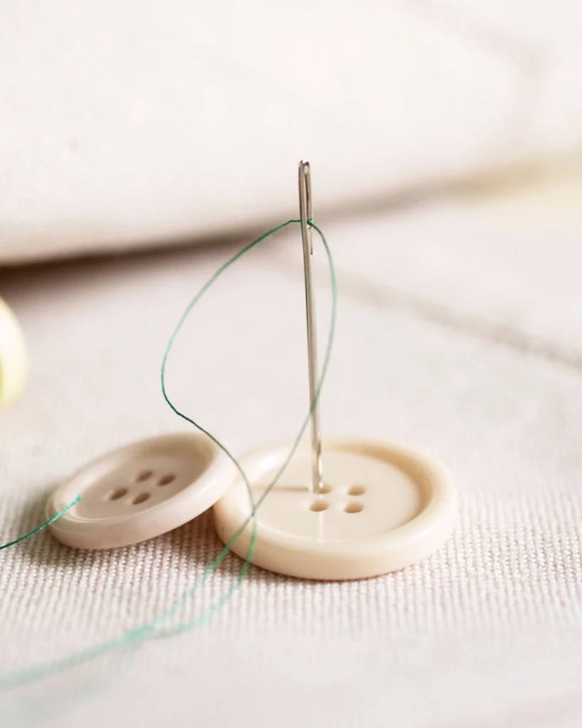 aguja con hilo verde costura botones blancos tela gruesa algodón crudo